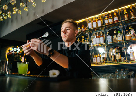Low-angle view of barman male squeezing out piece of orange peel with straw and decorating cocktail, standing behind bar counter in modern nightclub with dark interior. Low-angle view of barman male squeezing out piece of orange peel with straw and decorating cocktail, standing behind bar counter in modern nightclub with dark interior. 89151635