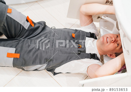 A male plumber repairs siphon under the sink in the bathroom. A male plumber repairs siphon under the sink in the bathroom. 89153653
