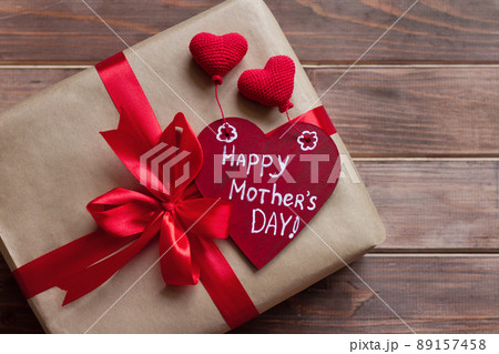 gift in brown craft paper with red ribbon with bow and wooden red heart with white inscription happy mother's day, knitted hearts on old brown table gift in brown craft paper with red ribbon with bow and wooden red heart with white inscription happy mother's day, knitted hearts on old brown table 89157458
