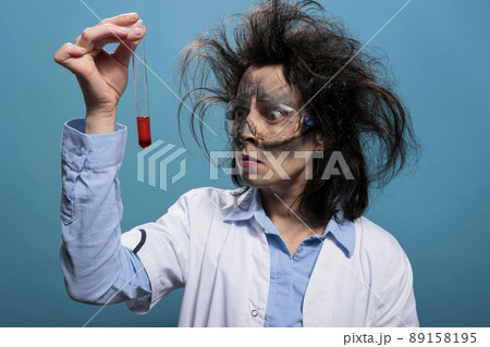 Mad worried chemist with messy hair and dirty face frowning at flask filled with red liquid substance sample. Insane anxious looking scientist analyzing chemistry glass tube filled with blood sample. 89158195