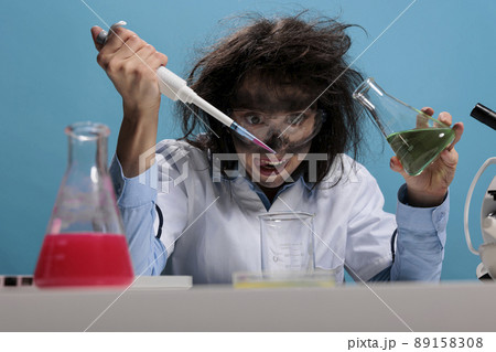 Foolish silly female lab worker with pipette and glass jar mixing toxic chemical compounds to create new formula. Crazy mad scientist mixing liquid substances while grinning dreadful. Foolish silly female lab worker with pipette and glass jar mixing toxic chemical compounds to create new formula. Crazy mad scientist mixing liquid substances while grinning dreadful. 89158308
