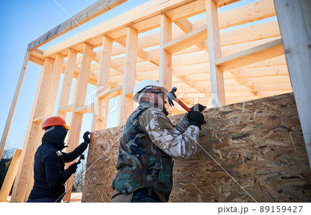Carpenters hammering nail into OSB panel on the wall of future cottage. Men workers building wooden frame house. Carpentry and construction concept. 89159427