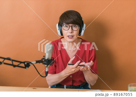 Mature woman making podcast recording for her online show. Attractive business woman using headphones front of microphone for a radio broadcast Mature woman making podcast recording for her online show. Attractive business woman using headphones front of microphone for a radio broadcast 89167055