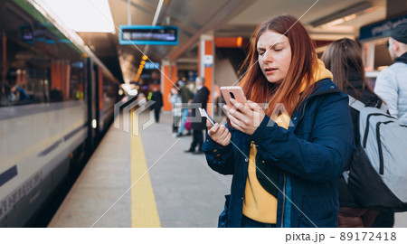 Young redhhead woman waiting on station platform with backpack on background electric train using smart phone. Railroad transport concept, Traveler. Invasion in Ukraine. 89172418