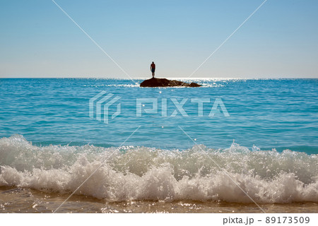Waves breaking on the sandy beach. The man stands on a small rocky island by the sea and getting ready to jump into the water. 89173509