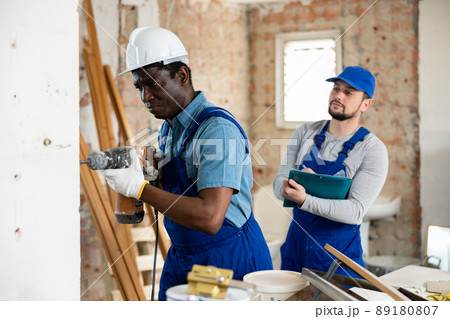 Builder drills a wall with a hammer drill, and his colleague makes notes 89180807