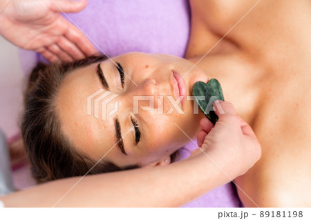 Relaxing massage. European woman getting quartz guache face massage in spa salon, side view Relaxing massage. European woman getting quartz guache face massage in spa salon, side view 89181198
