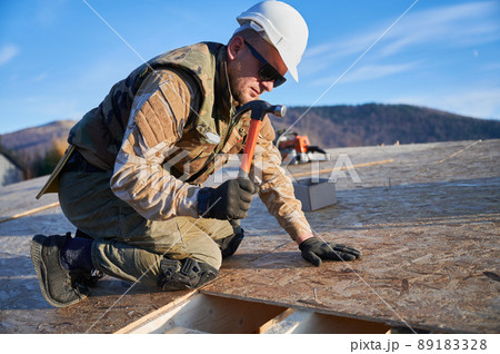 Carpenter hammering nail into OSB panel on the roof top of future cottage. Man worker building wooden frame house. Carpentry and construction concept. Carpenter hammering nail into OSB panel on the roof top of future cottage. Man worker building wooden frame house. Carpentry and construction concept. 89183328
