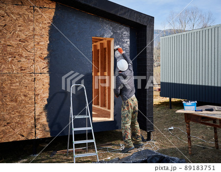 Male painter using paint roller, doing exterior paint work in a black color. Man worker building wooden frame house. Carpentry and construction concept. 89183751