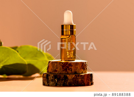 Cosmetic glass dropper bottle on wooden podium with oil, serum or fruit peeling in the sunlight. Green eucalyptus, soft focus. Beauty product presentation, natural background. Front. Mockup concept  89188388