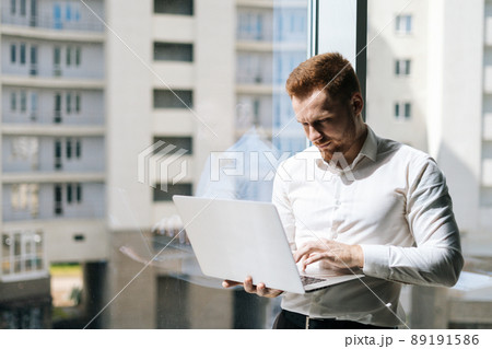 Medium shot portrait of focused young man working typing on laptop holding with one hand standing in modern office room near window, in sunny summer day on background of buildings. 89191586