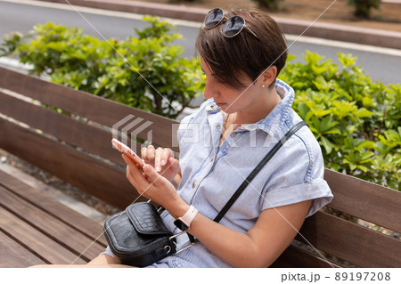 Young woman sitting on a bench in a park and typing on a phone. Young woman sitting on a bench in a park and typing on a phone. 89197208