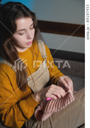 Attractive young woman knits woolen clothes while sitting on the sofa. Knitting hook. Home hobby production of things from natural wool Attractive young woman knits woolen clothes while sitting on the sofa. Knitting hook. Home hobby production of things from natural wool 89197455