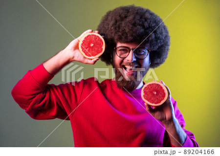 Portrait of hipster man with Afro hairstyle holding half slice grapefruit and smiling to camera, healthy raw fresh vitamin food concept. Indoor studio shot isolated on colorful neon light background. 89204166