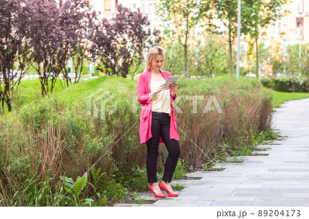 Full length portrait of happy beautiful young successful blogger businesswoman in elegance style standing on green park, holding tablet with toothy smile and typing message. Outdoor 89204173