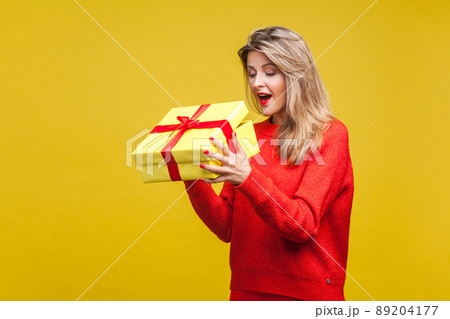 Portrait of surprised beautiful blonde woman with red lipstick in bright casual sweater, looking inside gift box with amazement, unboxing present. indoor studio shot isolated on yellow background Portrait of surprised beautiful blonde woman with red lipstick in bright casual sweater, looking inside gift box with amazement, unboxing present. indoor studio shot isolated on yellow background 89204177
