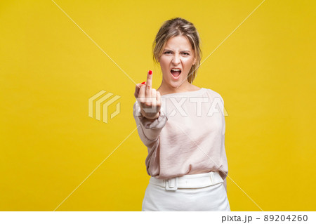 Portrait of angry dissatisfied young woman with fair hair in casual beige blouse standing, showing middle finger f you, looking at camera with hate. indoor studio shot isolated on yellow background 89204260