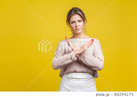 No more, this is the end. Portrait of serious stubborn young woman with fair hair in casual blouse standing showing closed gesture with crossed hands. indoor studio shot isolated on yellow background 89204275