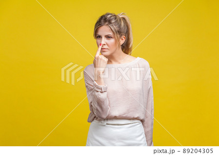 You are liar. Portrait of dishonest young woman with fair hair in casual beige blouse standing, touching her nose with finger, showing lie gesture. indoor studio shot isolated on yellow background 89204305