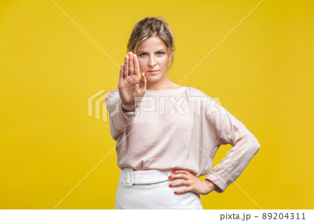 Portrait of confident serious young woman with fair hair in casual beige blouse standing, looking at camera, showing stop gesture, warning expression. indoor studio shot isolated on yellow background Portrait of confident serious young woman with fair hair in casual beige blouse standing, looking at camera, showing stop gesture, warning expression. indoor studio shot isolated on yellow background 89204311