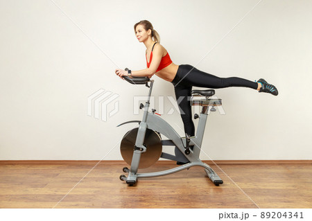 Side view portrait of slim attractive woman standing on exercise bike with raised leg, cardio workout, wearing sports tights and red top. Indoor studio shot on gray wall background. Side view portrait of slim attractive woman standing on exercise bike with raised leg, cardio workout, wearing sports tights and red top. Indoor studio shot on gray wall background. 89204341