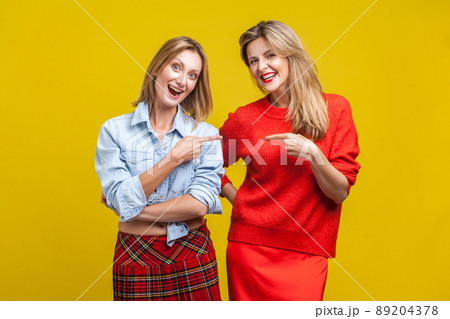 Best friends joking. Portrait of two happy women in casual clothes pointing and laughing at each other, looking at camera, carefree female friendship. indoor studio shot isolated on yellow background Best friends joking. Portrait of two happy women in casual clothes pointing and laughing at each other, looking at camera, carefree female friendship. indoor studio shot isolated on yellow background 89204378