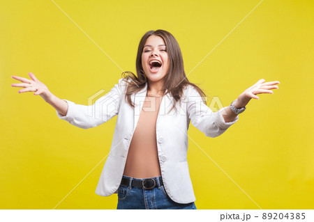 Portrait of delighted beautiful brunette woman in jacket and jeans raising hands in welcome gesture screaming for joy, excited to see you and going to hug. studio shot isolated on yellow background 89204385