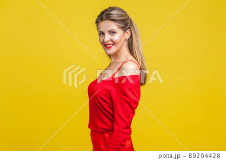 Half turn portrait of happy elegant young lady with neat ponytail hairstyle, bright makeup in red dress standing, looking at camera with toothy smile. indoor studio shot isolated on yellow background Half turn portrait of happy elegant young lady with neat ponytail hairstyle, bright makeup in red dress standing, looking at camera with toothy smile. indoor studio shot isolated on yellow background 89204428