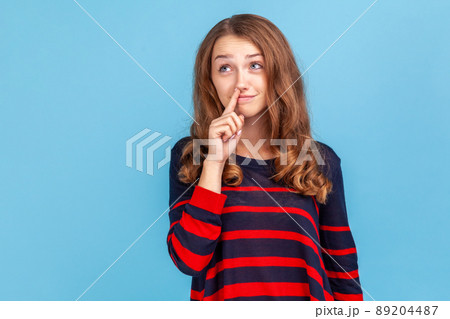 Childish woman wearing striped casual style sweater holding finger in her nose, uncultured bored girl having fun, bad manners, looking away. Indoor studio shot isolated on blue background. Childish woman wearing striped casual style sweater holding finger in her nose, uncultured bored girl having fun, bad manners, looking away. Indoor studio shot isolated on blue background. 89204487