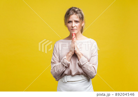 Portrait of worried young woman with fair hair in casual blouse standing, holding hands in prayer and looking at camera, asking help or forgiveness. indoor studio shot isolated on yellow background Portrait of worried young woman with fair hair in casual blouse standing, holding hands in prayer and looking at camera, asking help or forgiveness. indoor studio shot isolated on yellow background 89204546