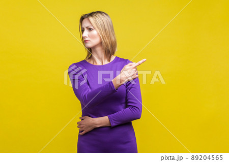 Portrait of angry bossy woman in elegant tight purple dress looking aside and pointing finger another way, asking to leave, dissatisfied expression. indoor studio shot isolated on yellow 89204565