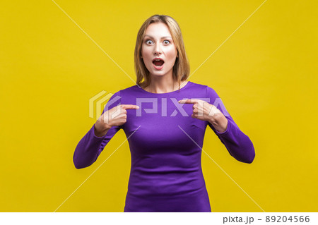 Portrait of amazed woman in elegant purple dress looking at camera with open mouth and pointing at herself, shocked about own success. indoor studio shot isolated on yellow background 89204566