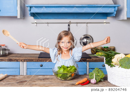 Lovely child girl joyfully raising her hands, holding wooden spoon and smiling looking at camera, happy to cook salad of fresh green vegetables, preparing healthy breakfast, vegetarian food, vitamins Lovely child girl joyfully raising her hands, holding wooden spoon and smiling looking at camera, happy to cook salad of fresh green vegetables, preparing healthy breakfast, vegetarian food, vitamins 89204746