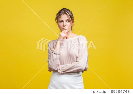 Portrait of pensive or confused young woman with blonde hair in casual beige blouse standing, holding her chin and thinking over problem solution. indoor studio shot isolated on yellow background Portrait of pensive or confused young woman with blonde hair in casual beige blouse standing, holding her chin and thinking over problem solution. indoor studio shot isolated on yellow background 89204748