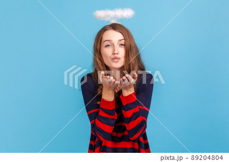 Kind woman wearing striped casual style sweater and nimb over head, sending air kiss over palms, expressing fondness, romantic feelings. Indoor studio shot isolated on blue background. 89204804