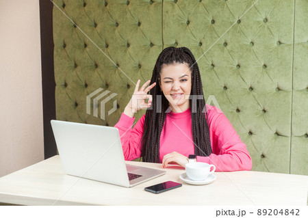 Portrait of positive funny young woman with black dreadlocks hairstyle in pink blouse sitting in cafe and showing ok sign with hand, winking, looking at camera and toothy smile. Indoor, lifestyle Portrait of positive funny young woman with black dreadlocks hairstyle in pink blouse sitting in cafe and showing ok sign with hand, winking, looking at camera and toothy smile. Indoor, lifestyle 89204842