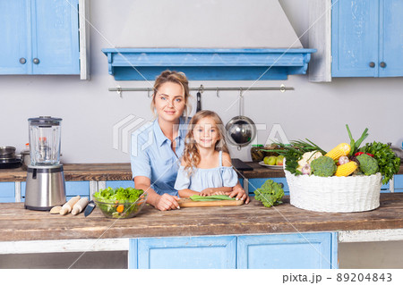 Young woman and lovely girl sitting in modern kitchen, looking at camera, mother and daughter cooking salad together, vegetarian food, fresh green vegetables and blender on table, healthy nutrition 89204843