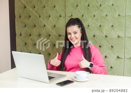 Portrait of attractive positive young girl freelancer with black dreadlocks hairstyle in pink blouse sitting in cafe and working on laptop with toothy smile and showing thumb up, looking at camera. 89204882
