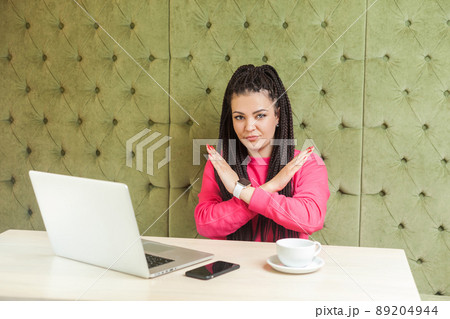 Warning aggressive young girl with black dreadlocks in pink blouse are sitting in cafe and working on laptop, showing crossing raised arms like stop or end gesture, looking at camera. Indoor Warning aggressive young girl with black dreadlocks in pink blouse are sitting in cafe and working on laptop, showing crossing raised arms like stop or end gesture, looking at camera. Indoor 89204944
