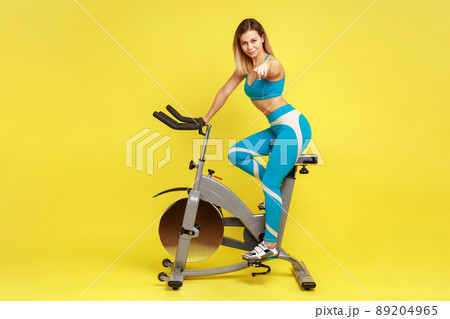Full length portrait of blonde woman coach on riding bike simulator, pointing to camera, choosing you for personal workout, wearing blue sportswear. Indoor studio shot isolated on yellow background. 89204965