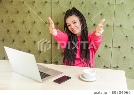 Portrait of lovely romantic young girl freelancer with black dreadlocks hairstyle are sitting alone in cafe and are ready hugging you with raised arms, toothy smile. Indoor, looking at camera Portrait of lovely romantic young girl freelancer with black dreadlocks hairstyle are sitting alone in cafe and are ready hugging you with raised arms, toothy smile. Indoor, looking at camera 89204966