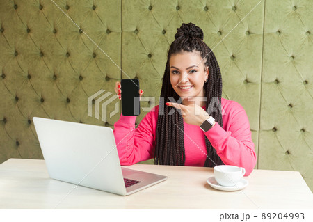 Portrait of beautiful satisfied happy young businesswoman with black dreadlocks in pink blouse is sitting in cafe, holding phone and pointing finger, looking at camera with toothy smile. Indoor 89204993