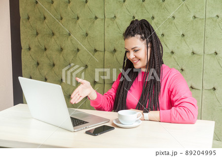 Portrait of happy young woman with black dreadlocks hairstyle in pink blouse sitting in and talking to webcamera at laptop and greeting with toothy smile, giving hand to online deal. indoor, lifestyle 89204995