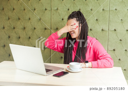 Portrait of emotional scared or shocked young businesswoman with black dreadlocks hairstyle in pink blouse sitting in cafe and covering her eyes and do not want to look at laptop screen. Indoor 89205180