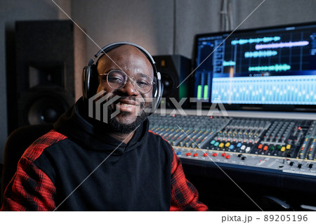 Portrait of cheerful African American music producer wearing headphones and eyeglasses sitting in recording studio smiling at camera 89205196