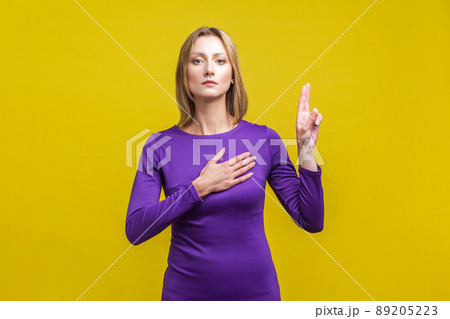 Portrait of honest citizen, young woman in elegant tight purple dress standing with hand on chest and fingers up, making loyalty promise. indoor studio shot isolated on yellow background 89205223