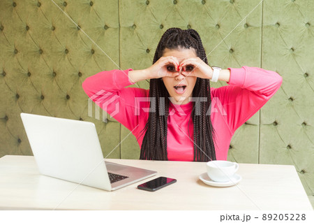 Suprised young woman with black dreadlocks in pink blouse sitting in cafe, holding her hands near eyes like making binoculas looking and try to find someone with open mouth. indoor Suprised young woman with black dreadlocks in pink blouse sitting in cafe, holding her hands near eyes like making binoculas looking and try to find someone with open mouth. indoor 89205228