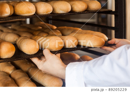 Closeup of hands of professional chef man in white uniform standing near shelves full with fresh bread and pulling baking tray for checking quality, Indoor, profession concept Closeup of hands of professional chef man in white uniform standing near shelves full with fresh bread and pulling baking tray for checking quality, Indoor, profession concept 89205229