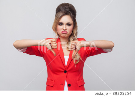 Portrait of sad dissatisfied beautiful business lady with hairstyle and makeup in red fancy blazer, standing with thubms down and looking at camera. indoor studio shot, isolated on grey background. Portrait of sad dissatisfied beautiful business lady with hairstyle and makeup in red fancy blazer, standing with thubms down and looking at camera. indoor studio shot, isolated on grey background. 89205254