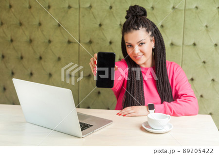 Portrait of successful beautiful young woman with black dreadlocks in pink blouse is sitting, holding phone empty screen and giving you, looking at camera with toothy smile. Indoor, business concept Portrait of successful beautiful young woman with black dreadlocks in pink blouse is sitting, holding phone empty screen and giving you, looking at camera with toothy smile. Indoor, business concept 89205422